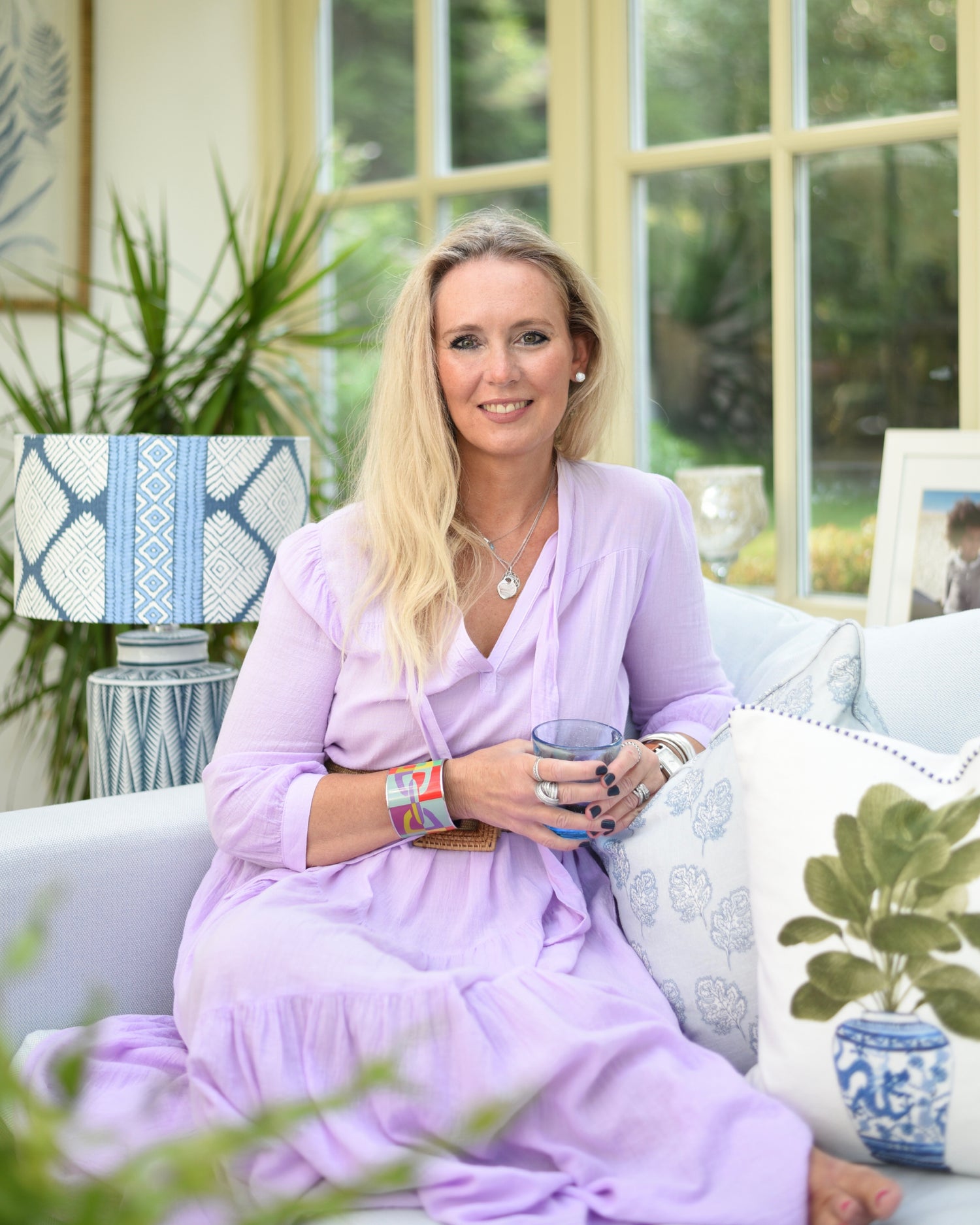 Woman in a purple outfit sitting on a couch holding a cup, with a decorative lamp and plants in the background.